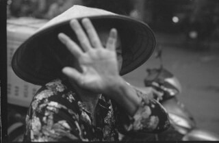 A person wearing an Asian conical hat obscures their face with their hand in this black and white image.