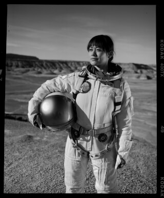A woman in a vintage astronaut suit stands in a desert landscape, holding her helmet and looking off into the distance.