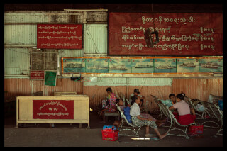 People sit and wait indoors in Burma, with weathered walls and local signs creating an authentic scene.