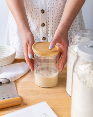A person prepares sourdough starter in a glass jar on a wooden table.
