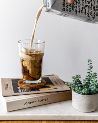 Creamy oat milk is poured into a glass of coffee on a stack of books.