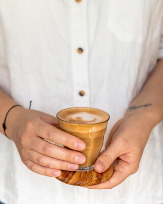 Hands hold a latte in a glass on a wooden coaster, a comforting and warm coffee break moment.