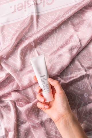 Hand holding a Glossier priming moisturizer tube against a pink patterned cloth.