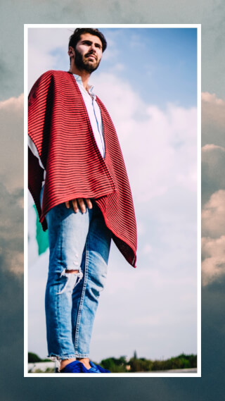 A bearded man in a red poncho and jeans stands against a cloudy sky.