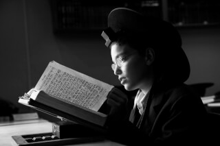 A boy wearing glasses and a kippah is deeply engrossed in reading an open book in black and white.
