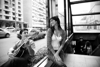 A street musician plays guitar for a woman sitting near a subway entrance in a black and white urban scene.