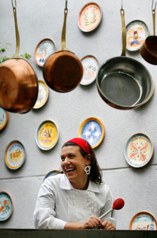 A laughing chef in a white coat stands amidst hanging copper pots and decorative plates.
