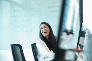 A happy businesswoman laughs while sitting in an office environment during a meeting, with a whiteboard in the background.