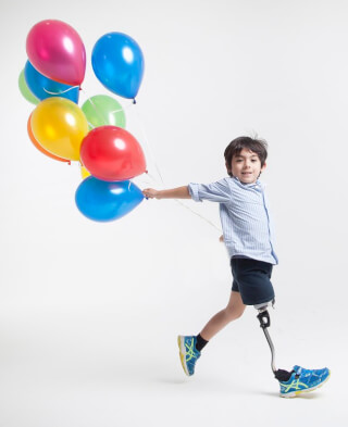 A happy young boy with a prosthetic leg runs while holding colorful balloons in a studio setting.