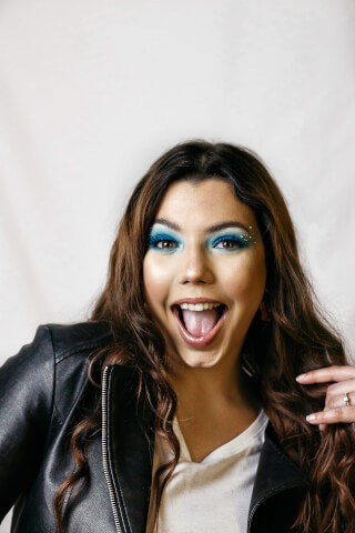 A cheerful woman with vibrant blue eyeshadow and a leather jacket smiles with an open mouth.