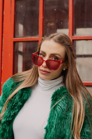 A woman with red sunglasses and a green faux fur coat poses against a red door.