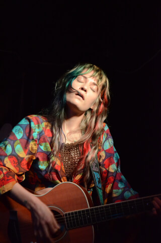A woman with colorful hair performs with an acoustic guitar in a close-up shot on a dimly lit stage.