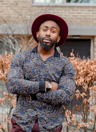 A stylish man poses with arms crossed wearing a hat and patterned shirt in an urban outdoor setting.