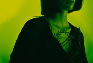 A woman in dark clothing with a leafy garland against a green background conveys a sense of mystery and natural beauty.