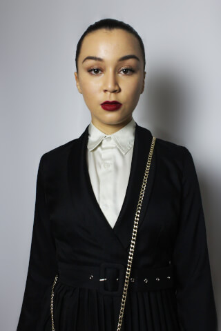 A woman with red lipstick wears a black suit jacket and white collared shirt in a studio setting.