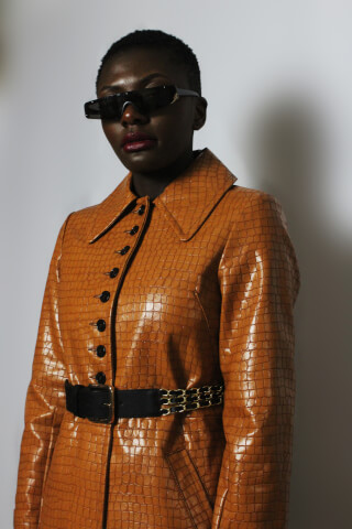 A confident woman poses in a stylish brown crocodile-print jacket with a belt and sunglasses.
