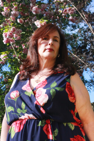 A mature woman in a floral dress poses outdoors surrounded by blooming trees in natural light.