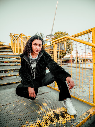 A young woman with curly hair squats on metal stairs in an urban setting, wearing casual clothing.