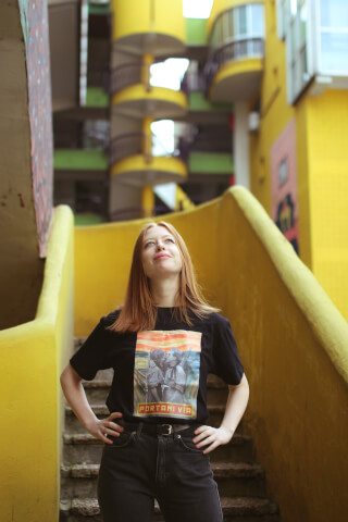 A redhead woman poses on yellow stairs in an urban setting, wearing a graphic tee.