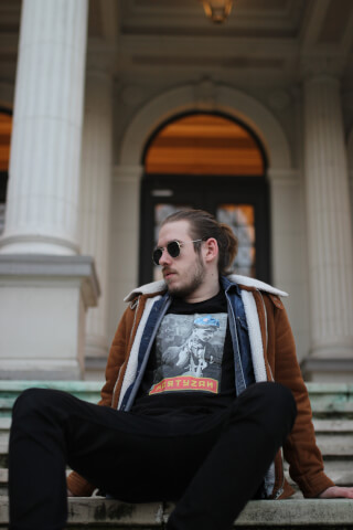 Man sits on steps wearing sunglasses, a jacket, and a t-shirt with a graphic design.