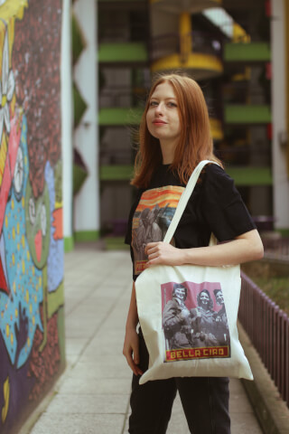 A redhead woman in trendy streetwear poses with a tote bag against an urban mural backdrop.