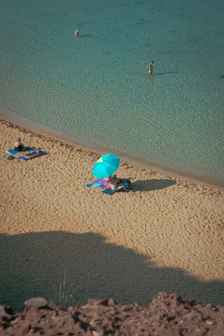 A sunny beach scene with people relaxing under an umbrella and swimming in clear turquoise water.
