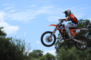 A motocross rider soars through the air on an orange KTM bike against a blue sky with clouds.