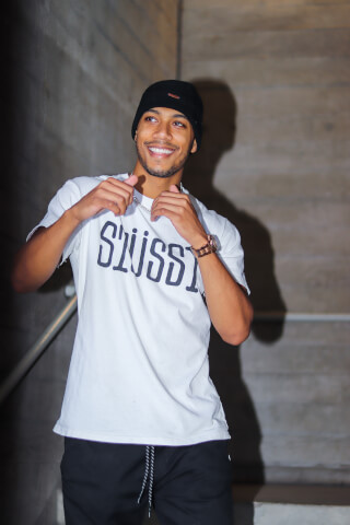 A smiling man in a beanie and graphic tee poses in an urban stairwell setting, exuding confidence and casual style.