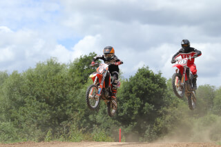 Two motocross riders are airborne on a dirt track, racing towards the finish line in an action-packed sports event.