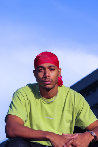 A confident young man poses with a red durag and green t-shirt against a blue sky.