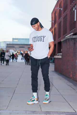 A smiling man wearing a Stussy t-shirt checks his phone on a city street with a brick building in the background.