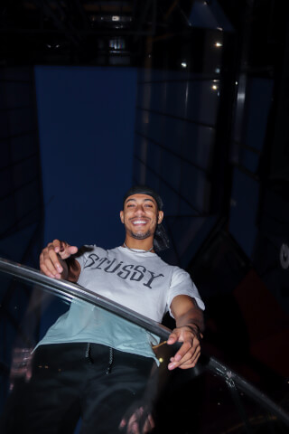 A smiling man in a Stussy t-shirt poses in a modern urban setting with cool tones.