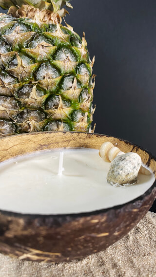 A tropical candle in a coconut shell sits beside a pineapple, perfect for relaxation and beach-themed decor.