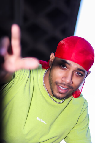 A stylish man in a red durag and green shirt smiles and reaches towards the camera.