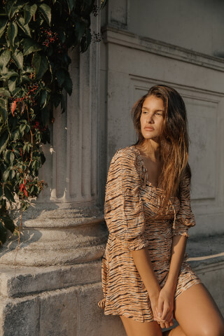 A brunette woman in a tiger-striped dress poses outdoors, bathed in soft light.