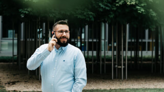 A bearded man with glasses has a phone call outside in a park setting.