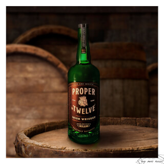 A bottle of Proper Twelve Irish Whiskey sits on a wooden barrel in a dimly lit setting.