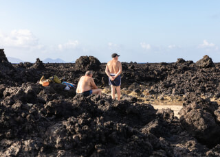 Two shirtless men in swim trunks relax on a rocky beach with black lava rocks on a sunny day.