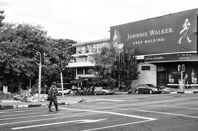 A pedestrian crosses a street in an urban area with a large Johnnie Walker advertisement billboard visible.
