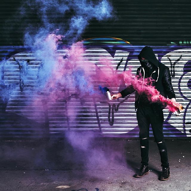 A hooded figure with a mask creates a colorful smoke display with spray cans in an urban setting.