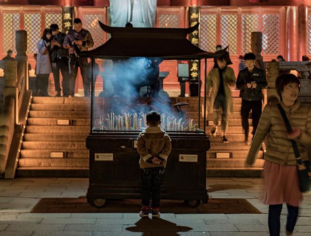 A boy stands before a smoking incense burner at a temple, with people on steps in the background.