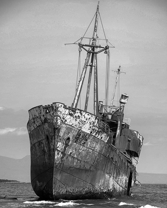 An abandoned, rusty shipwreck sits offshore under a cloudy sky, showcasing its weathered, monochrome beauty.