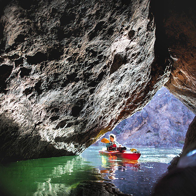 A kayaker paddles a red kayak through a calm river cave surrounded by rock formations.