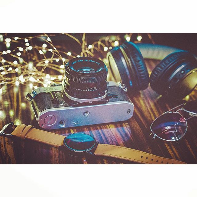 Vintage camera, headphones, watch, and sunglasses arranged on a wooden table with string lights.