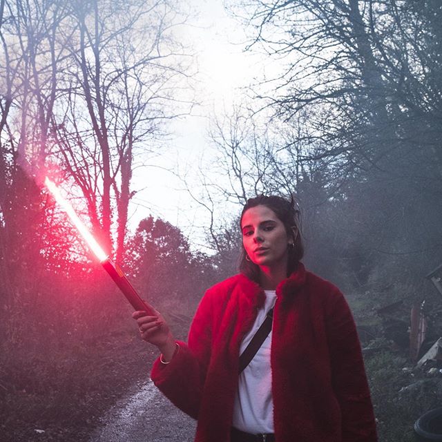 A woman in a red coat poses with a red lightsaber in a misty forest setting.