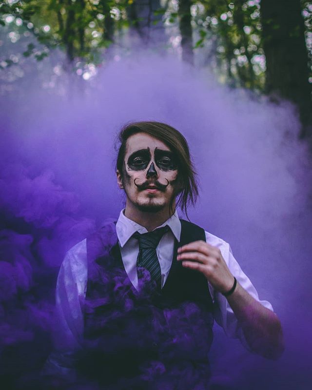 A person in a vest with skeleton face makeup stands in a forest surrounded by purple smoke.