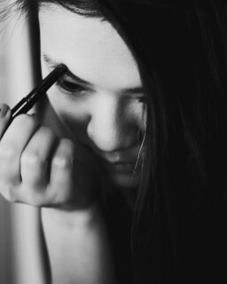 A woman carefully applies makeup to her eyebrow in a close-up, monochrome studio shot.