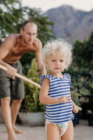 A toddler in a striped shirt plays outside while her father tends to the garden.