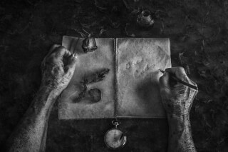 Monochrome image of hands writing 'The End' in a vintage book with dried flowers and a pocket watch.