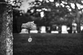 A close-up of an elderly hand holding a pocket watch in a cemetery, evoking a sense of time and mortality.